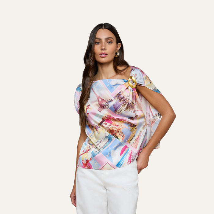 A woman wearing the Driselle Scarf Top, a colorful, patterned short-sleeve blouse, paired with a gold brooch and white pants, poses with one hand in her pocket against a plain background.