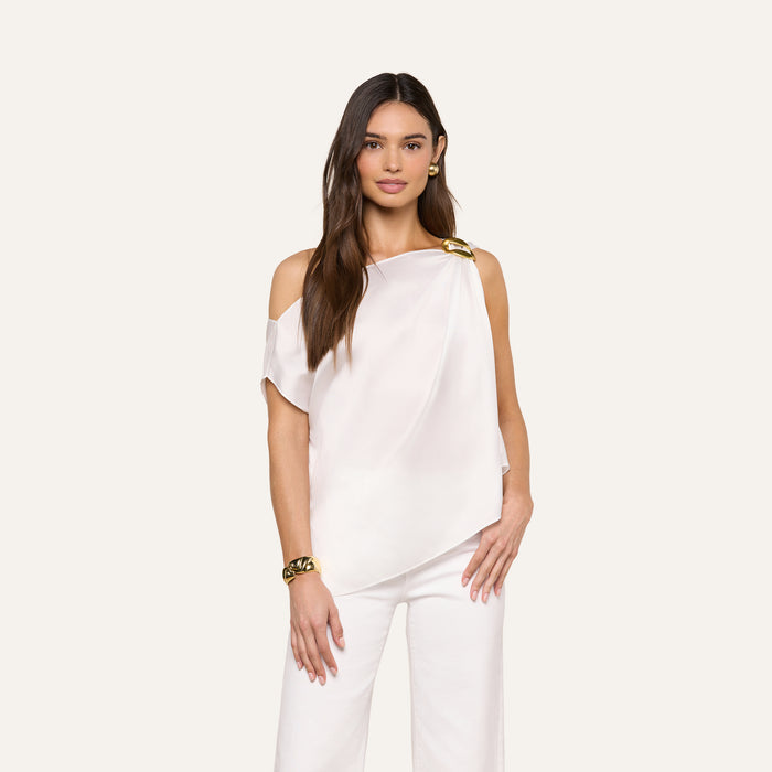 A woman with long brown hair models the Driselle Scarf Top—an off-the-shoulder satin piece with gold hardware—paired with matching white pants, gold earrings, and a bracelet against a beige background.