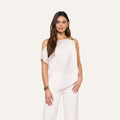 A woman with long brown hair models the Driselle Scarf Top—an off-the-shoulder satin piece with gold hardware—paired with matching white pants, gold earrings, and a bracelet against a beige background.