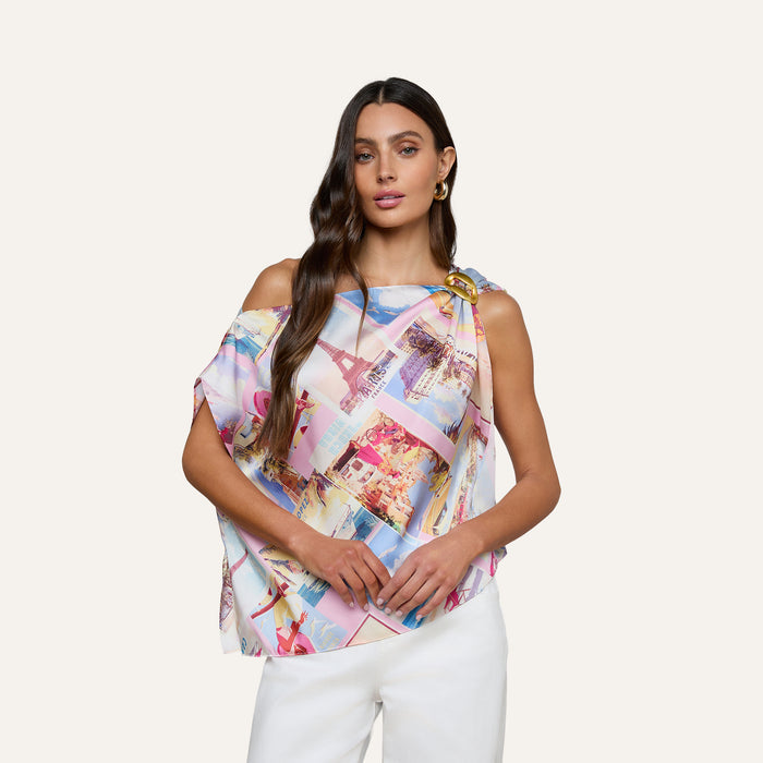 A woman with long brown hair models the Driselle Scarf Top, featuring a vibrant postcard print and an asymmetrical design, paired with white pants against a plain light background—making it an ideal pick for vacation wear.