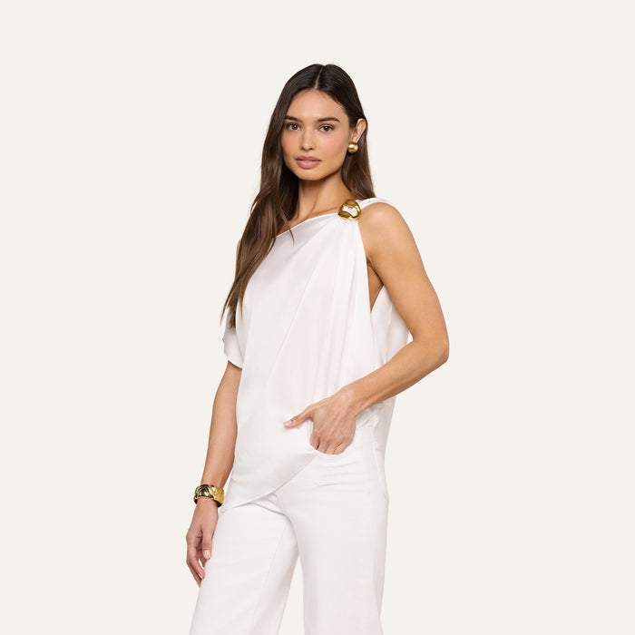 A woman with long brown hair poses against a beige background wearing the Driselle Scarf Top, a sleeveless white satin piece with gold accents on the shoulders, paired with white pants and gold jewelry.