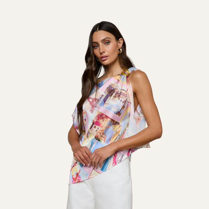 A woman with long dark hair wears the Driselle Scarf Top, featuring a colorful postcard print and a gold buckle detail, paired with white pants, standing against a plain light background.