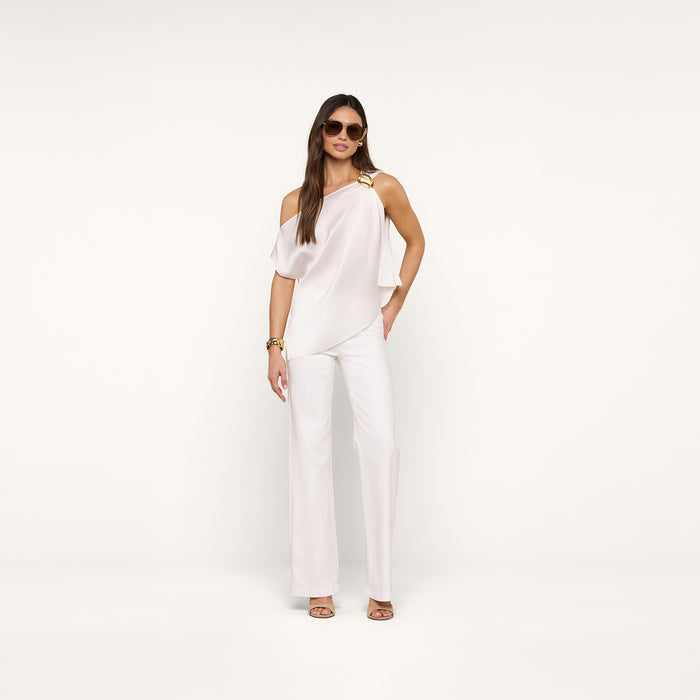 A woman poses confidently against a beige background wearing sunglasses, the Driselle Scarf Top with a gold buckle, white wide-leg pants, gold heels, and a gold bracelet.