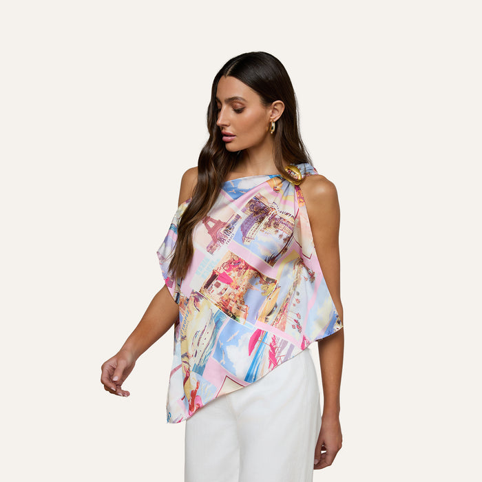 A woman wearing the Driselle Scarf Top—a colorful, asymmetrical postcard-print design—paired with white pants, stands against a plain light background, looking down and to the side.
