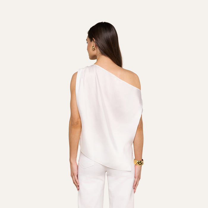 A woman with long brown hair stands facing away, wearing the Driselle Scarf Top in white satin and matching pants, accessorized with gold earrings and a bracelet against a beige background.