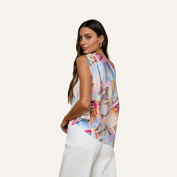 A woman with long brown hair wears the Driselle Scarf Top—a colorful postcard print sleeveless blouse—paired with white pants, looking over her shoulder against a plain light background.
