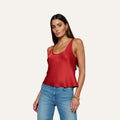 A woman with long dark hair wears the Eddie Tank, a bias-cut satin poppy red sleeveless top with a scoop neckline, paired with blue jeans and accessorized with gold hoop earrings and a gold bracelet, standing against a plain light background.