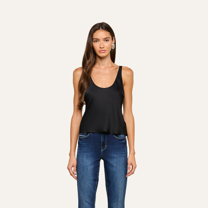 A woman with long brown hair stands facing forward against a plain light background, wearing the Eddie Tank—a black sleeveless top with a scoop neckline—paired with blue jeans.