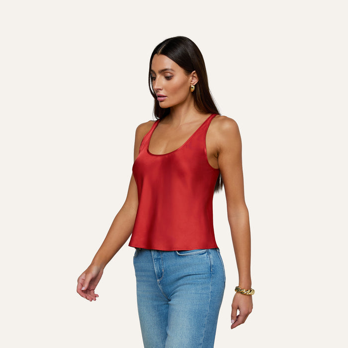 A woman with long brown hair wears the Eddie Tank, a sleeveless poppy red bias-cut satin top with a scoop neckline, paired with blue jeans. She accessorizes with gold hoop earrings and a gold bracelet, standing against a plain light background.