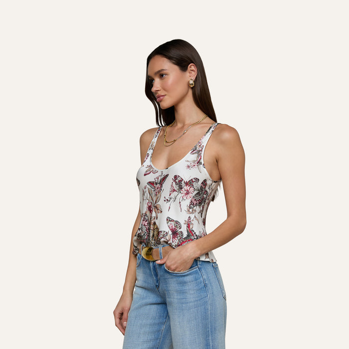 A woman wearing the Eddie Tank—a floral scoop-neck tank top—paired with blue jeans stands against a plain background, facing slightly to the left. She has long brown hair and accessorizes with earrings and a necklace.