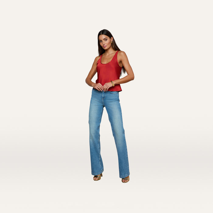 A woman stands confidently against a plain white background, wearing the Eddie Tank in poppy red—a sleeveless top with a scoop neckline—paired with light blue jeans and sandals.