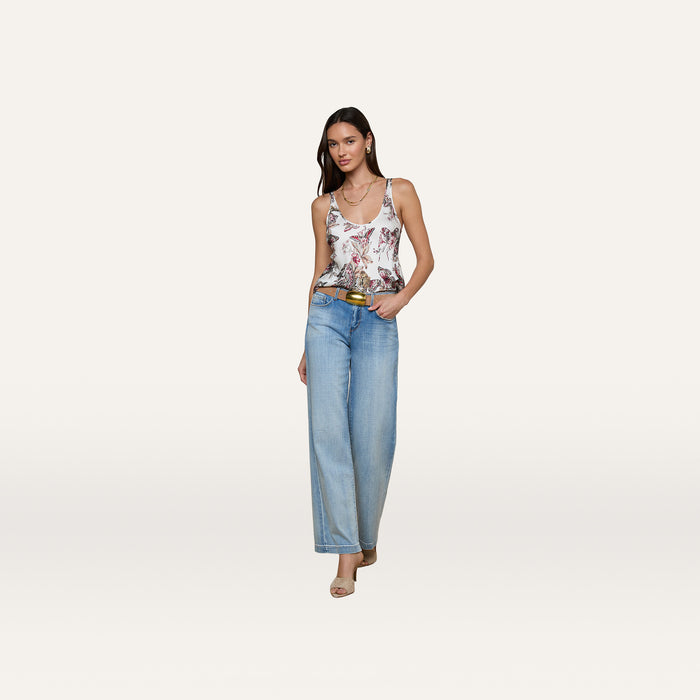 A woman wearing the Eddie Tank, a white floral tank top with a scoop neckline, styled with light blue wide-legged jeans, beige heels, and a belt poses against a plain white background.