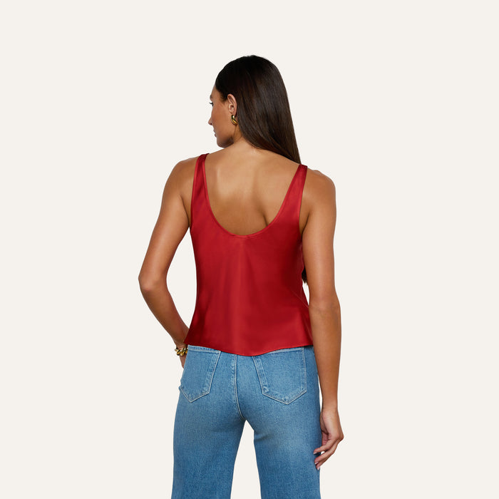 A woman with long brown hair, wearing the Eddie Tank—a bias-cut satin top in poppy red—paired with blue jeans, stands facing away from the camera against a plain light background.
