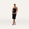 A woman wears the Eira Midi Dress, a fitted black slip dress with adjustable straps, paired with black heeled sandals as she stands against a plain white background.
