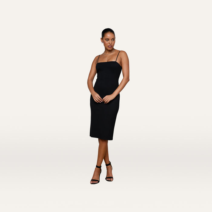 A woman wears the Eira Midi Dress, a fitted black slip dress with adjustable straps, paired with black heeled sandals as she stands against a plain white background.