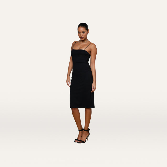 Wearing the Eira Midi Dress, a woman poses against a plain white background, looking at the camera with one hand by her side. The black knee-length slip dress features adjustable straps and is paired with black heels.