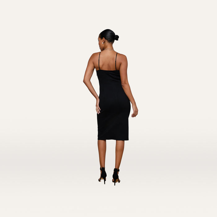 A woman with her hair in a bun wears the Eira Midi Dress—a fitted, sleeveless slip dress crafted from super stretch ponte—in black, paired with black heels. She stands with her back to the camera against a plain white background.