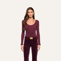 Woman with long brown hair, wearing the Elara Scoopneck Bodysuit—a fitted long-sleeve burgundy top crafted from stretch cotton modal—and high-waisted burgundy pants with a large rectangular belt, stands facing forward against a plain light background.