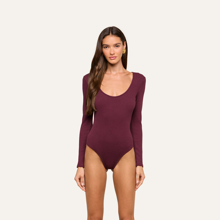 A woman with long brown hair is wearing the Elara Scoopneck Bodysuit in deep burgundy, which features a body-contouring fit made from stretch cotton modal. She is standing against a plain, light background and looking straight at the camera with a neutral expression.