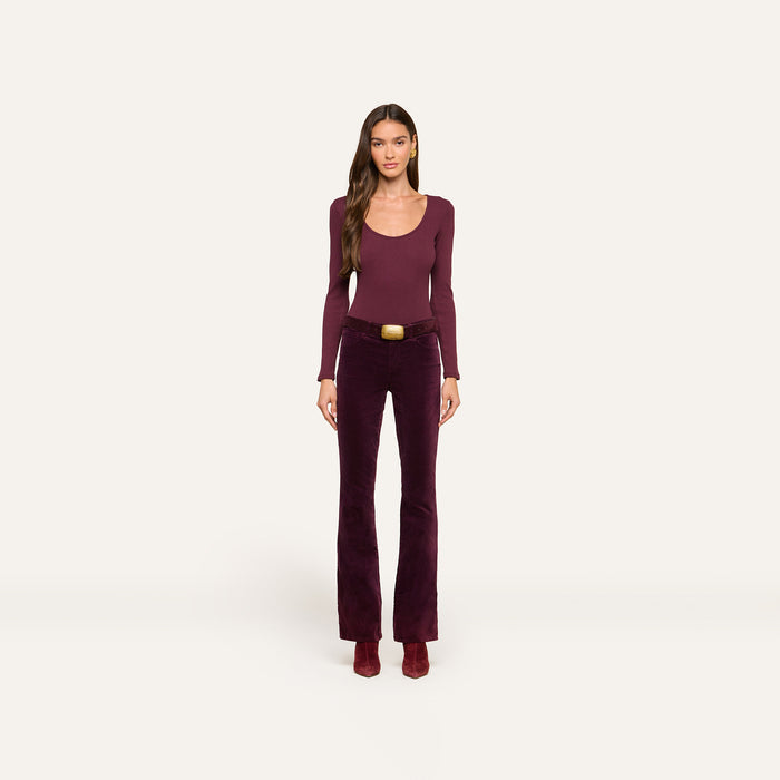 A woman stands facing forward against a plain white background, wearing a long-sleeve burgundy Elara Scoopneck Bodysuit with a body-contouring fit, matching flared pants with a belt, and burgundy boots.