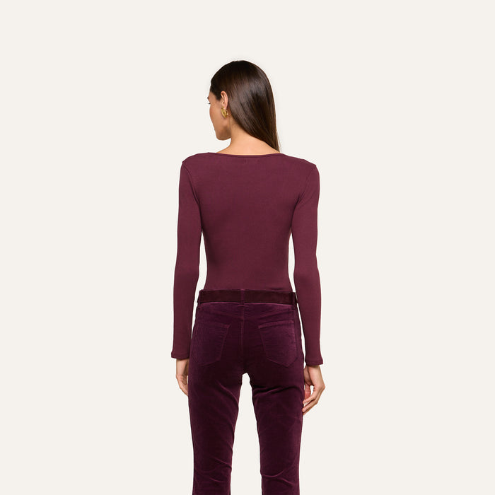 A woman stands with her back to the camera, wearing the Elara Scoopneck Bodysuit in maroon—crafted from stretch cotton modal—and high-waisted velvet pants in a coordinating shade, set against a simple light background. The look emphasizes the Elara’s sleek, body-contouring fit.