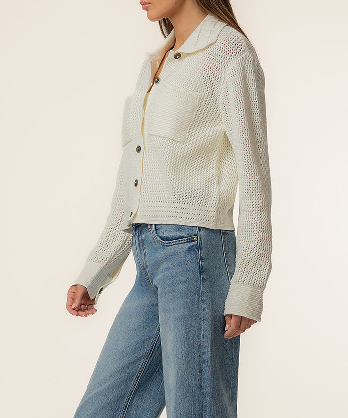 Woman wearing cream knit sweater jacket with button front and textured weave, styled with blue jeans.