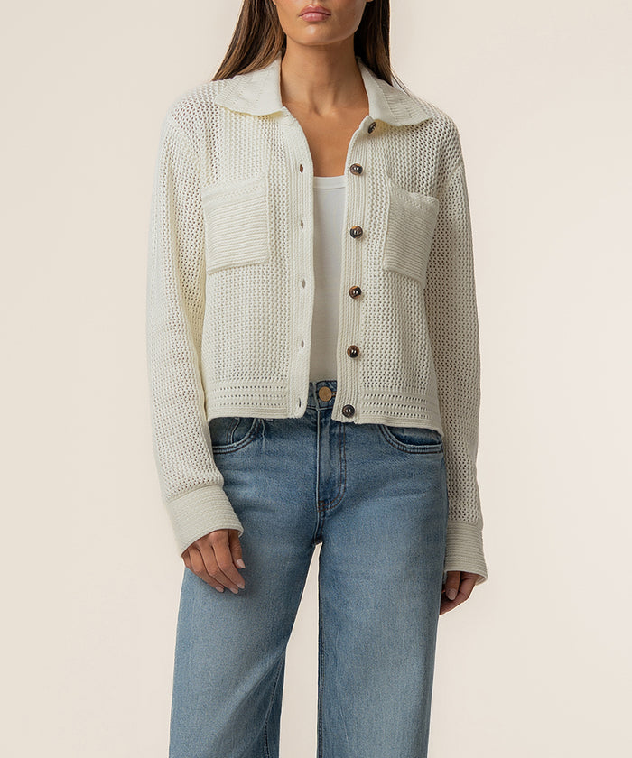 Cream knit sweater jacket with textured panels, collar, and brown button front closure worn over white top and blue jeans.