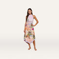 Barefoot, a woman poses against a plain white background in the Elise Cover-Up Dress—a sleeveless, halter midi dress with a handkerchief hem, featuring a vibrant scenic postcard print of a tropical city and palm trees.