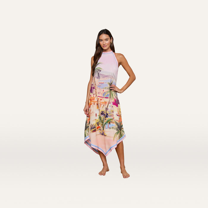 Barefoot, a woman poses against a plain white background in the Elise Cover-Up Dress—a sleeveless, halter midi dress with a handkerchief hem, featuring a vibrant scenic postcard print of a tropical city and palm trees.