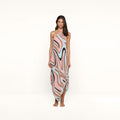 A woman stands barefoot against a white background, wearing the Elise Cover-Up Dress—a halter midi dress crafted from water-safe crepe and showcasing an abstract swirl print in pink, beige, blue, and black tones.