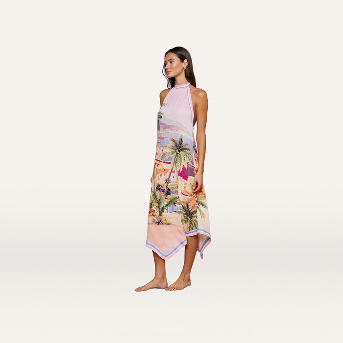 A woman stands barefoot wearing the Elise Cover-Up Dress, a water-safe crepe halter midi dress adorned with a vibrant scenic postcard print of palm trees and buildings, set against a plain white background.