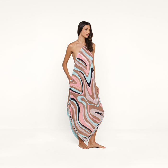 A woman stands barefoot wearing the Elise Cover-Up Dress, a halter midi dress featuring a colorful abstract swirl print in pink, blue, brown, and black on a plain white background.