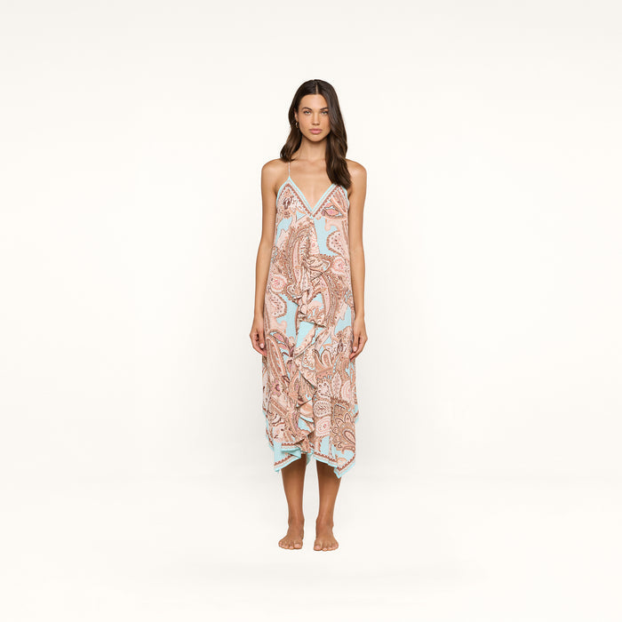 A woman stands barefoot against a white background, wearing the Elise Cover-Up Dress—a halter midi dress crafted from water-safe crepe. The dress features a paisley print in pastel colors, thin straps, and a handkerchief hem.