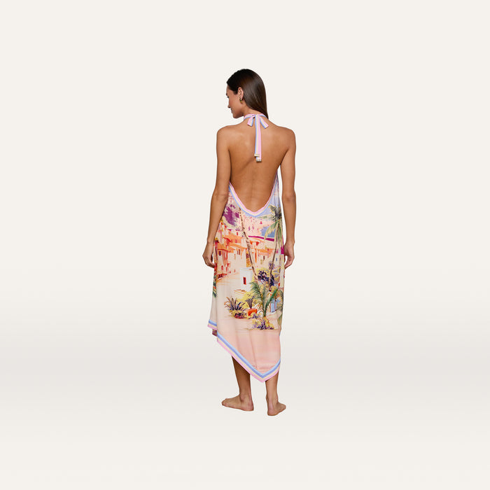 A woman stands barefoot, shown from the back, wearing the Elise Cover-Up Dress—a water-safe crepe halter midi dress with an open back and a colorful scenic postcard print depicting buildings and palm trees. The background is plain white.