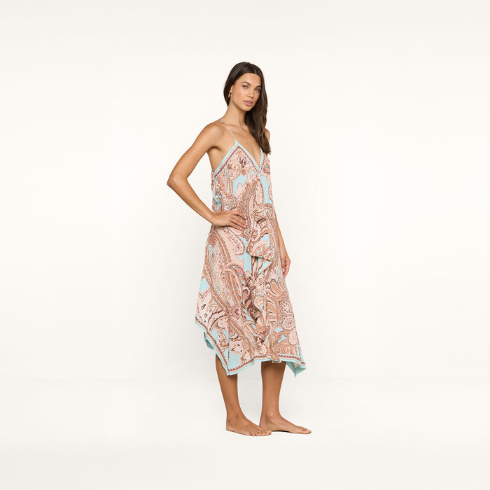 A woman stands barefoot against a plain white background, wearing the Elise Cover-Up Dress—a sleeveless, flowy halter midi dress featuring a paisley print in soft pastel colors.