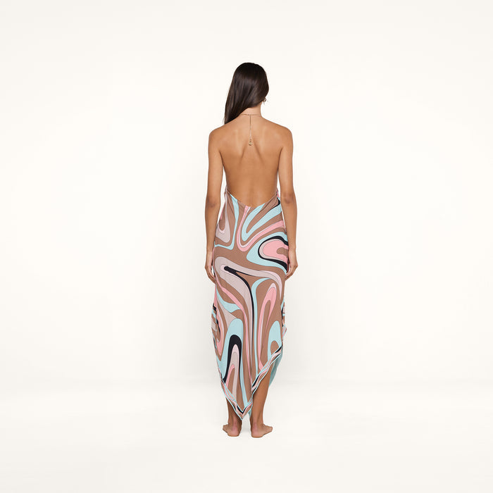 Barefoot and facing away, a woman with long brown hair wears the Elise Cover-Up Dress—a halter midi dress crafted from water-safe crepe. The backless design showcases an abstract swirl print in pink, blue, black, and white on a white background.