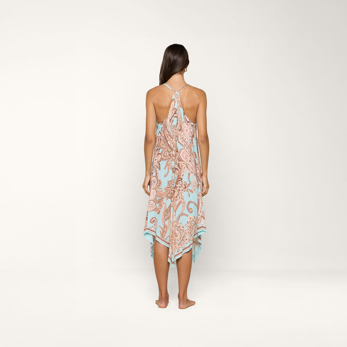 A woman stands barefoot, facing away, wearing the Elise Cover-Up Dress in a light blue and brown paisley print with a halter neckline and an uneven hem, set against a plain white background.