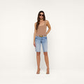 A woman stands against a plain white background wearing sunglasses, a tan sleeveless top, heeled sandals, and the Elise Jean Bermuda Short—light blue low-rise shorts crafted from all-cotton denim with a relaxed fit.