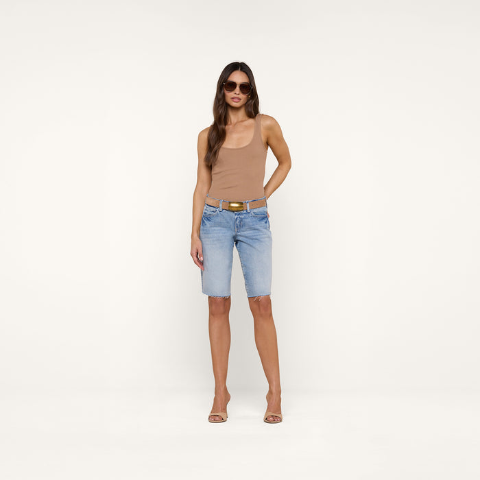A woman stands against a plain white background wearing sunglasses, a tan sleeveless top, heeled sandals, and the Elise Jean Bermuda Short—light blue low-rise shorts crafted from all-cotton denim with a relaxed fit.