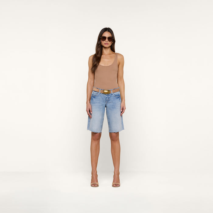 A woman stands against a plain white background wearing sunglasses, a tan tank top, and the Elise Jean Bermuda Short—light blue low-rise shorts with a relaxed fit crafted from all-cotton denim. She completes her look with heeled sandals.