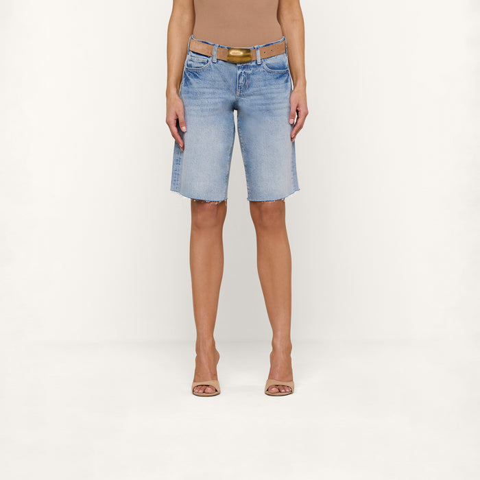 A person wearing a tan sleeveless top, a tan belt, open-toe heels, and the Elise Jean Bermuda Short—a relaxed fit, light blue, knee-length all-cotton denim short with a raw hem—stands against a plain white background.