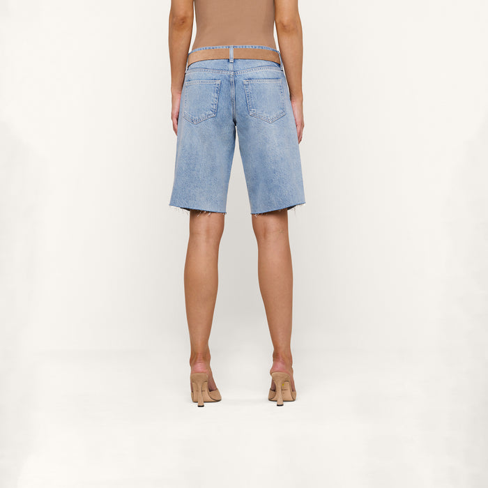A person is shown from the back against a plain white background, wearing a tan top, nude high heels, and the Elise Jean Bermuda Short—relaxed fit, all-cotton denim knee-length shorts in light blue.