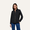 A woman wearing the Ellery Button-Down Shirt, a black stretch cotton blouse with gold buttons, paired with blue jeans stands against a plain light background, looking at the camera with one hand on her hip.