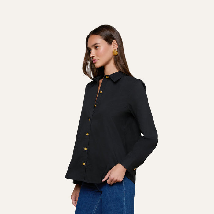 Wearing the Ellery Button-Down Shirt, a woman with long brown hair styles it with blue jeans and large round earrings, standing against a plain light background.