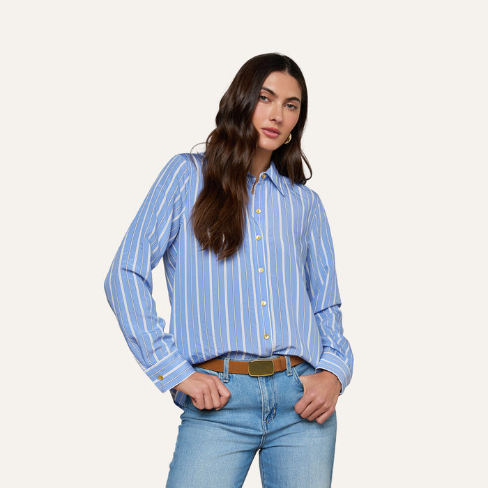 A woman with long dark hair stands against a plain background, hands in her pockets, wearing the Ellery Poplin Shirt— a blue and white striped button-down, tucked into light blue jeans.