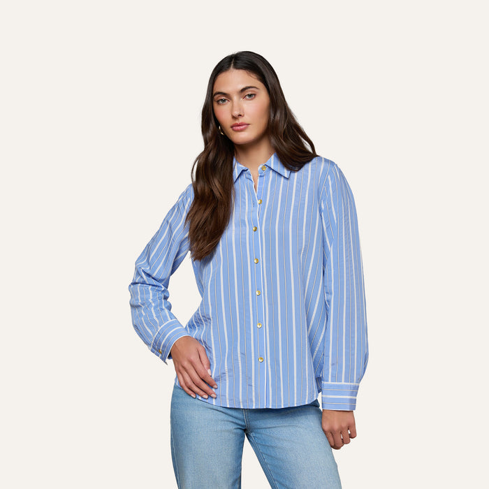 A woman models the Ellery Poplin Shirt—a blue and white striped, long-sleeve button-down—paired with light blue jeans against a plain off-white background.
