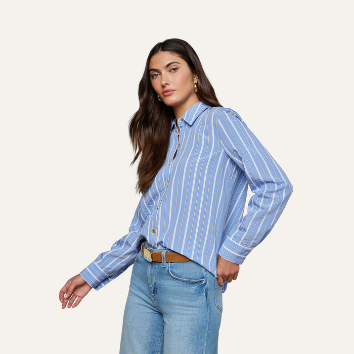 Sure Here’s the rewritten sentence using the product name: Woman with long brown hair wearing the Ellery Poplin Shirt—a blue and white striped button-down—paired with blue jeans and a brown belt, standing against a plain light background.