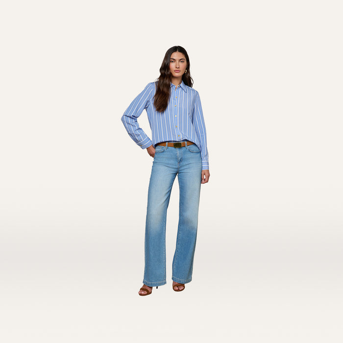 A woman wearing the Ellery Poplin Shirt—a relaxed fit blue striped button-down—styled with light blue wide-leg jeans, a brown belt, and brown heeled sandals stands against a plain white background.