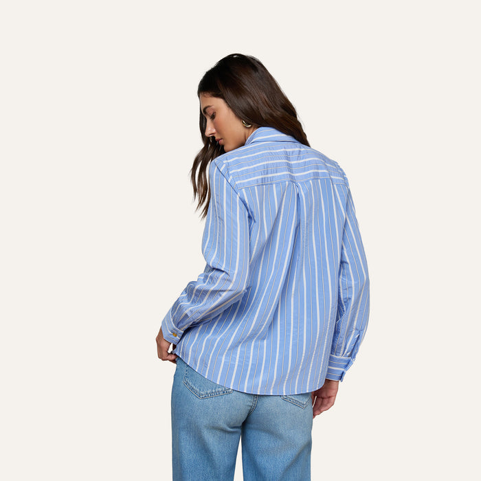 A woman with long brown hair, wearing the Ellery Poplin Shirt in a relaxed fit with stripes and light blue jeans, stands with her back to the camera against a plain light background.