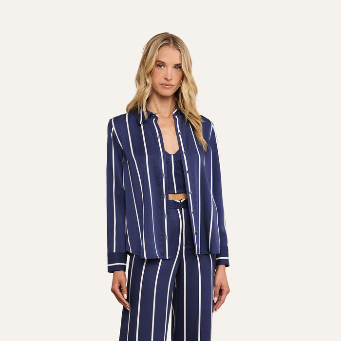 Wearing the Ellery Shirt—a lightweight Tencel navy blue button-down with white vertical stripes—and matching pants, the woman stands against a plain light background. The relaxed fit of the ensemble lends effortless style and comfort to her look.